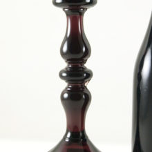In dark amethyst glass. Tall baluster stem. Stands 8 5/8" tall on a 4½" base. Candle hole is 1" wide with a drip rim that is 2 3/4" wide. The base is hollow and the stem is solid glass. Weighs 1 1/4 lbs.